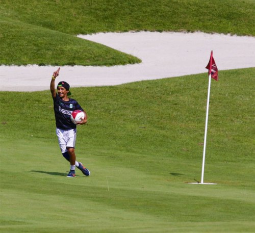 A young boy runs on a golf course, holding a soccer ball and pointing excitedly towards the flag.