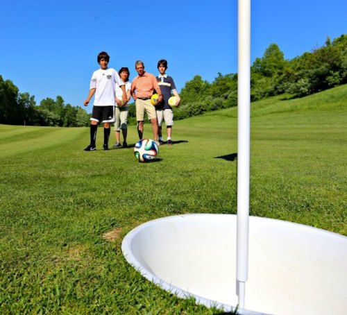A group of four people, including an older man, stands on a grassy field near a soccer ball and a goal hole.