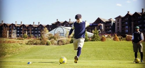 A golfer prepares to swing on a sunny day, with a resort in the background and another person watching nearby.