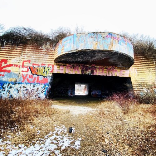 Abandoned structure with colorful graffiti, surrounded by overgrown grass and trees, under a cloudy sky.