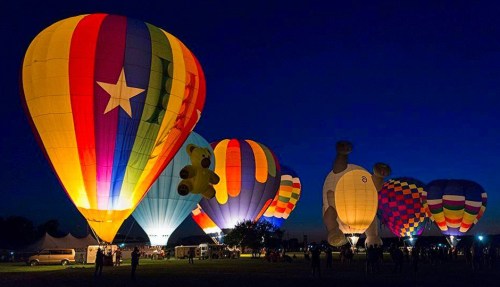Colorful hot air balloons illuminated at night, featuring various shapes and patterns against a dark blue sky.