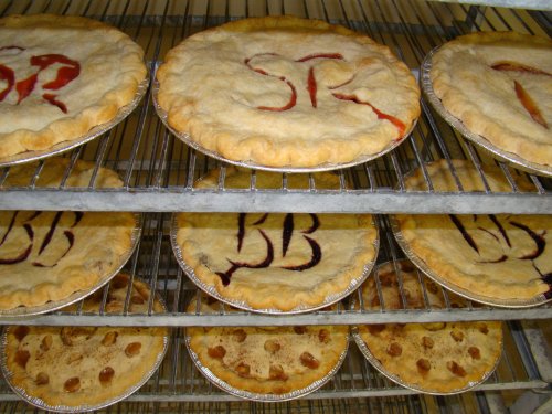 A rack of freshly baked pies with crusts marked by initials, some filled with fruit and others with a decorative pattern.