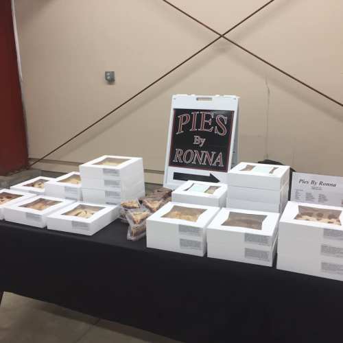 A display table with white boxes of pies and a sign reading "Pies By Ronna" in a market setting.