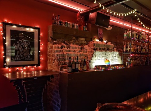 A cozy bar scene with a brick wall, colorful lights, and a framed artwork, showcasing shelves of bottles.