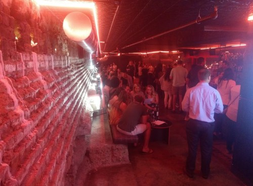 A dimly lit bar with red lighting, featuring a crowd of people socializing and a brick wall in the background.