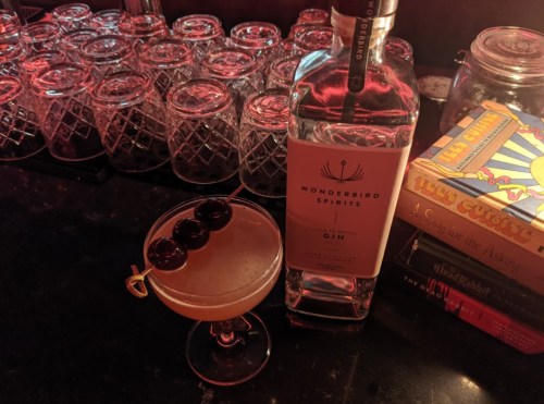 A cocktail with three cherries sits on a bar next to a bottle of Wonderbird gin and a stack of colorful books.