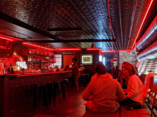 A dimly lit bar with red neon lights, featuring a long bar, patrons seated, and a stylish interior design.