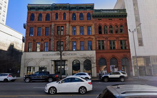 Historic brick buildings with large windows, featuring "Founder's Station" and "Masonry Building" signs, along a city street.