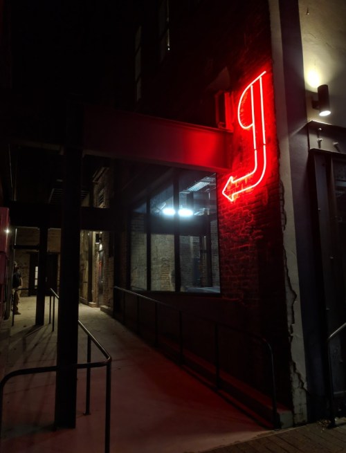 A dimly lit alley at night featuring a red neon "J" sign with an arrow pointing to the right.