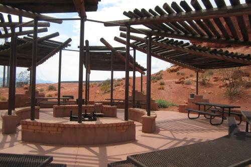 Outdoor gathering area with shaded structures, picnic tables, and a fire pit, surrounded by desert landscape.