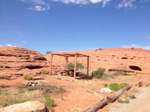 A wooden shelter with a picnic table in a desert landscape, surrounded by red rock formations and sparse vegetation.