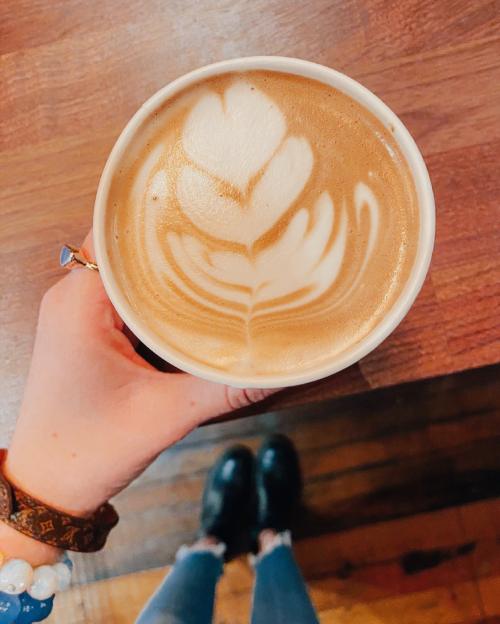 A hand holding a latte with a heart-shaped foam design, with a wooden table and feet in the background.