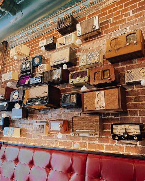 A wall adorned with various vintage radios, showcasing a mix of styles and colors against a brick background.