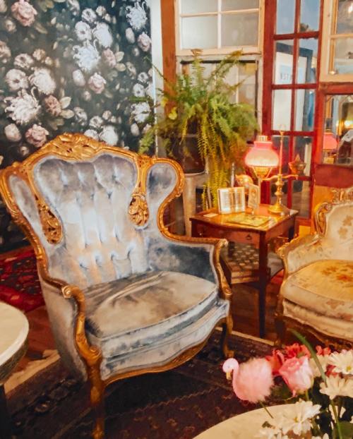 A vintage blue armchair with ornate gold detailing, surrounded by floral wallpaper, a small table, and decorative plants.