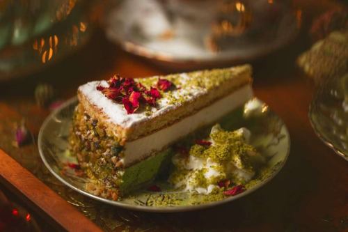 A slice of layered cake topped with rose petals and pistachios, served on a decorative plate with whipped cream.