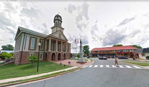 Historic building with a clock tower, statue in front, and a street with a pedestrian crossing nearby. Cloudy sky.