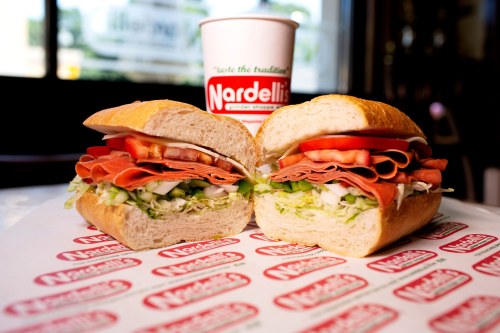 A freshly cut sandwich with layers of lettuce, tomatoes, and deli meats, alongside a cup from Nardelli's.