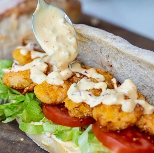 A close-up of a sandwich with crispy shrimp, lettuce, tomato, and a creamy sauce drizzled on top.