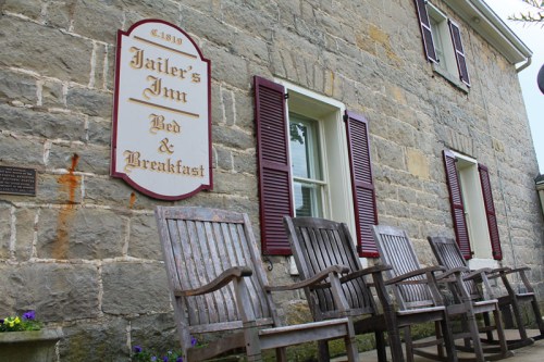 Sign for Jailer’s Inn Bed & Breakfast on a stone building, with wooden rocking chairs on the porch.