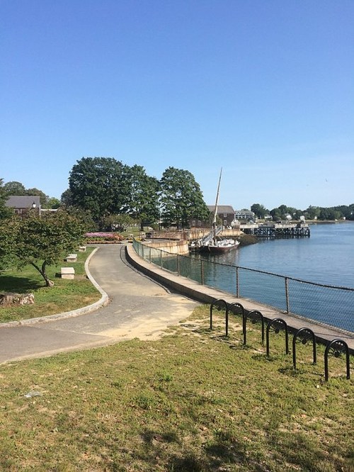 A scenic waterfront path lined with trees, leading to a docked boat and a clear blue sky.