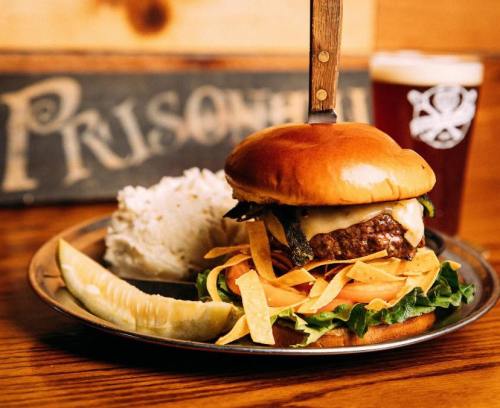 A juicy burger topped with cheese, lettuce, tomato, and tortilla strips, served with rice and a pickle on a metal plate.