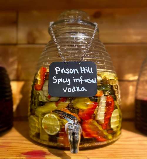 A glass dispenser filled with spicy infused vodka, featuring colorful peppers and limes, labeled "Prison Hill."
