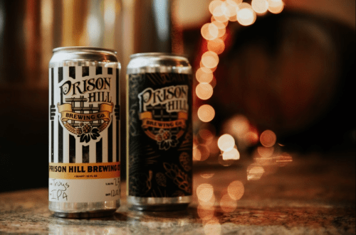 Two cans of Prison Hill Brewing Co. beer on a countertop, with blurred lights in the background.