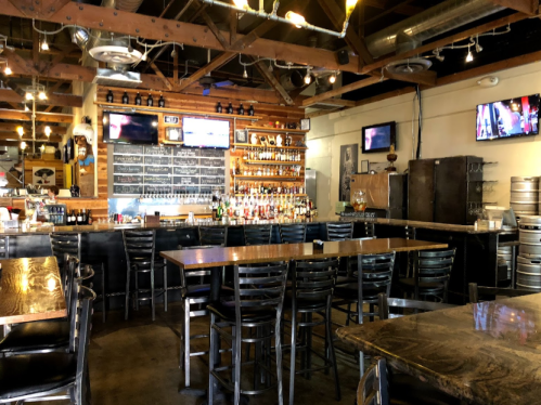 A rustic bar interior with wooden beams, a long table, and shelves stocked with various drinks. TVs are mounted on the walls.