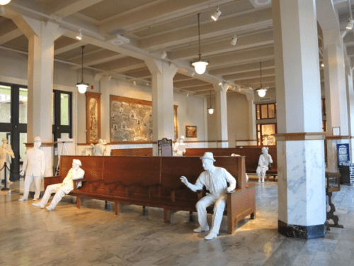 A spacious room with white-painted figures sitting on benches, surrounded by historical artwork and marble flooring.