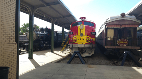 Two vintage trains are displayed under a shelter, one red and yellow, the other a classic steam locomotive.