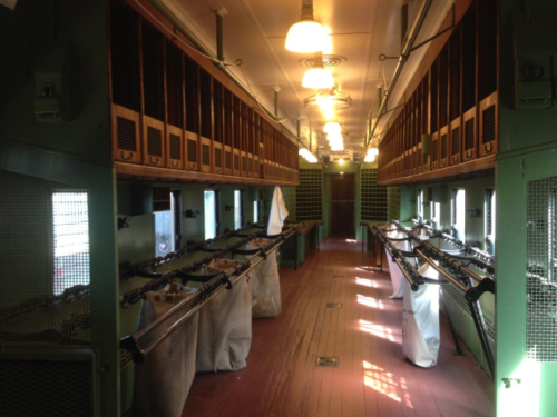 Interior of a vintage train car with green walls, wooden floors, and rows of storage compartments and bags.