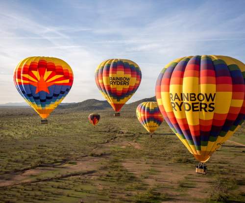Colorful hot air balloons with "RAINBOW RYDERS" logo flying over a scenic landscape under a clear sky.