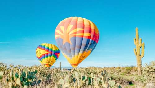 Two colorful hot air balloons float above a desert landscape with cacti under a clear blue sky.