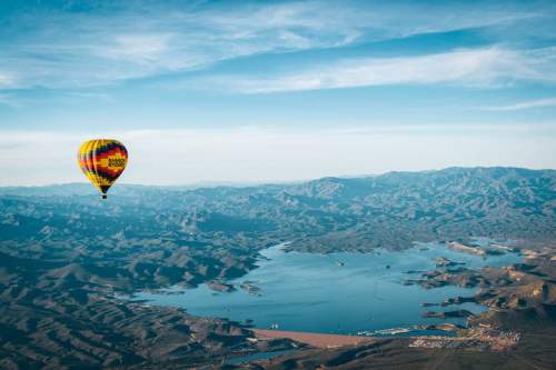 A colorful hot air balloon floats over a vast lake surrounded by mountains under a clear blue sky.