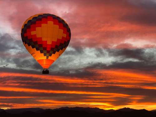 A colorful hot air balloon floats against a vibrant sunset sky with dramatic clouds and mountain silhouettes.