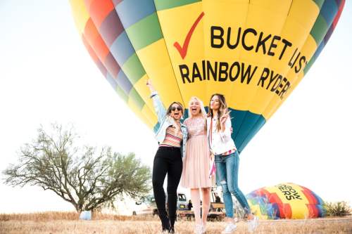 Three women joyfully pose in front of a colorful hot air balloon with "Bucket List" and "Rainbow Ryders" printed on it.