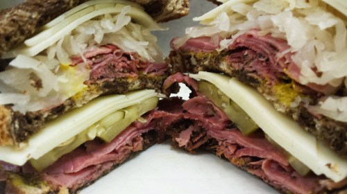 Close-up of a sliced sandwich with layers of pastrami, Swiss cheese, pickles, sauerkraut, and mustard on dark bread.