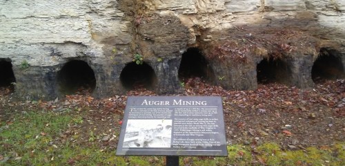 Sign about auger mining in front of several large, circular openings in a rocky wall, surrounded by fallen leaves.