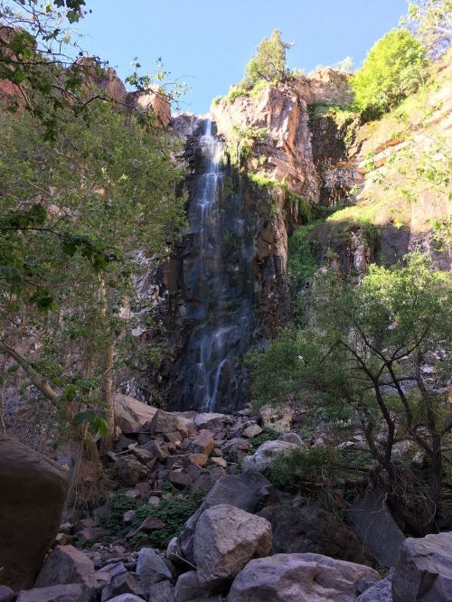 A cascading waterfall surrounded by rocky terrain and lush greenery under a clear blue sky.