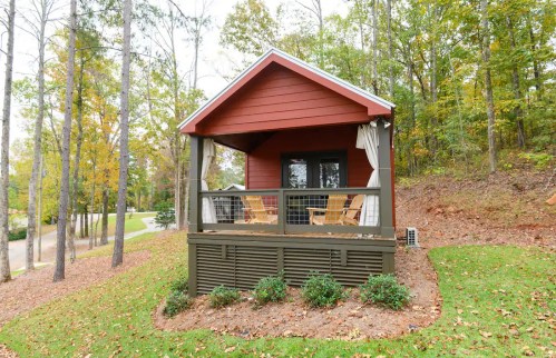 A cozy red cabin with a porch, surrounded by trees and greenery, set on a hillside.