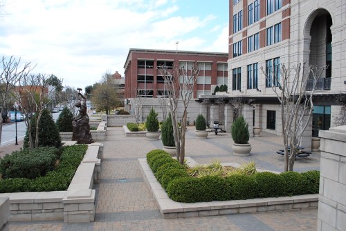 A landscaped plaza with trees and shrubs, adjacent to a modern building and a parking garage under a cloudy sky.