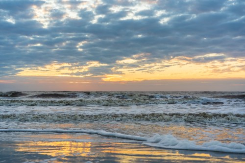 A serene beach scene at sunset, with waves gently lapping the shore under a cloudy sky.