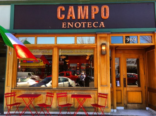 Exterior of Campo Enoteca restaurant with red tables, an Italian flag, and a welcoming wooden entrance.