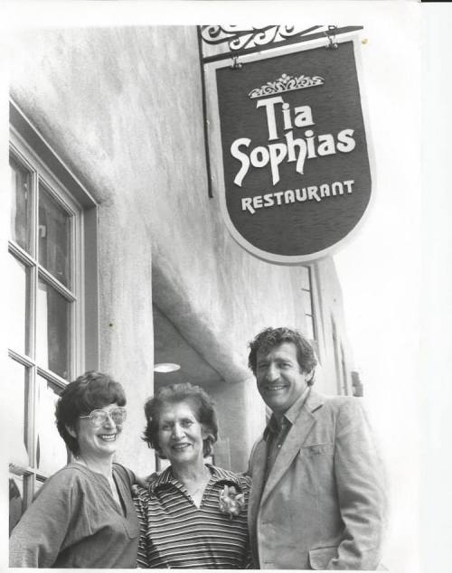 A black and white photo of three people smiling in front of a restaurant sign that reads "Tia Sophia's Restaurant."