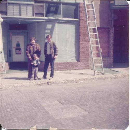 A woman, a man, and a child stand on a street in front of a building, with a ladder leaning against the wall.