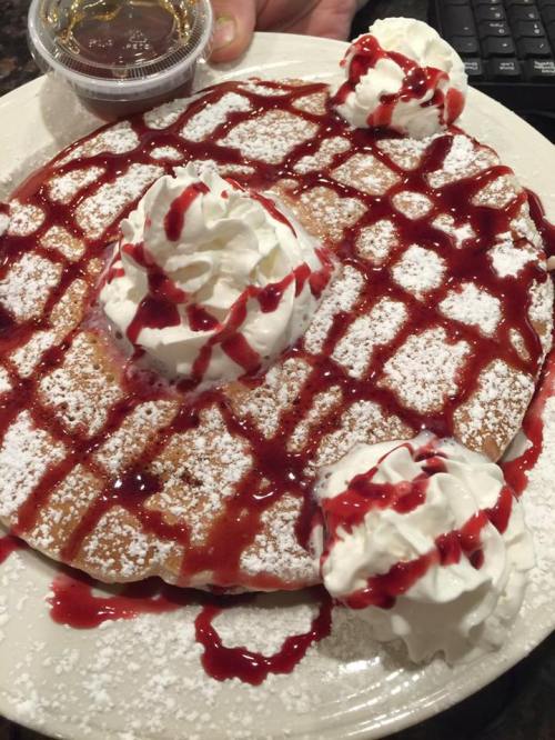 A large pancake topped with whipped cream, berry sauce, and powdered sugar, served with a small cup of syrup.