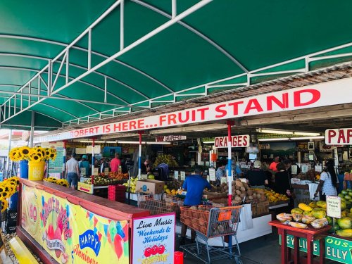 A bustling fruit stand with colorful produce, shoppers, and a sign reading "Robert is here... Fruit Stand." Sunflowers in view.