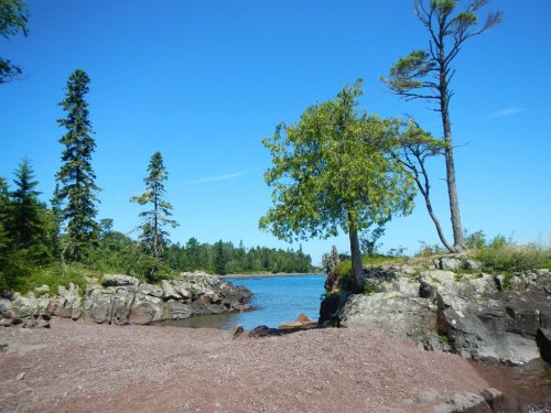A serene landscape featuring a rocky shore, trees, and a calm blue lake under a clear blue sky.