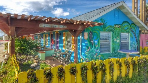 Colorful house with a mural, surrounded by greenery and a yellow picket fence, under a bright blue sky.