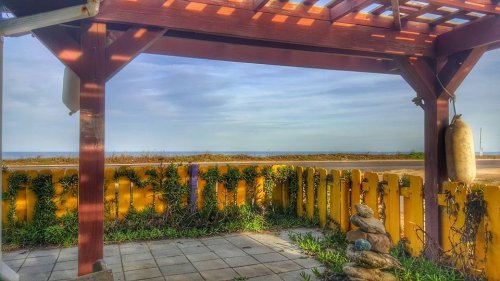 View from a wooden pergola overlooking a calm sea, with a yellow fence and stacked stones in the foreground.